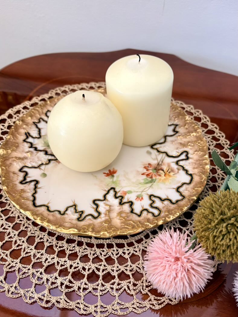 Two white candles on a decorative plate with a doily underneath, surrounded by flowers.