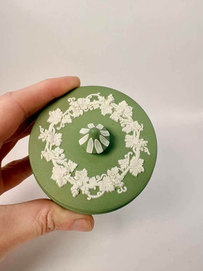 Green ceramic jar with white floral patterns held by a hand against a light gray background