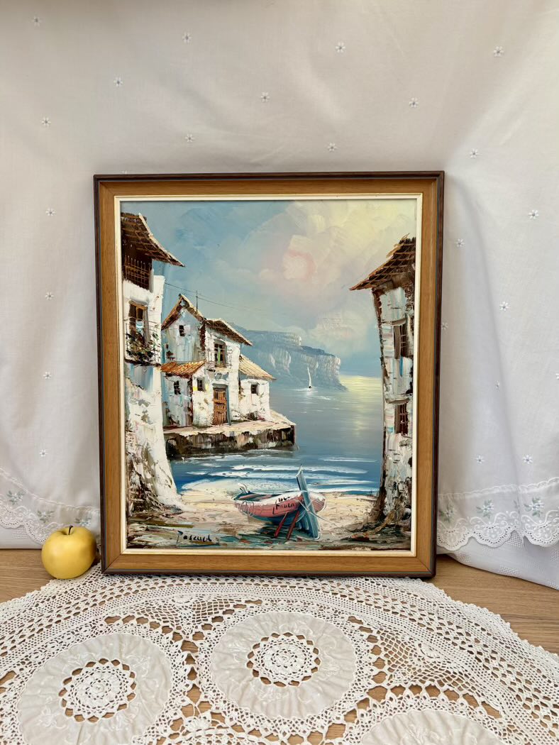 Framed painting of a coastal scene with houses and a boat on a lace tablecloth.
