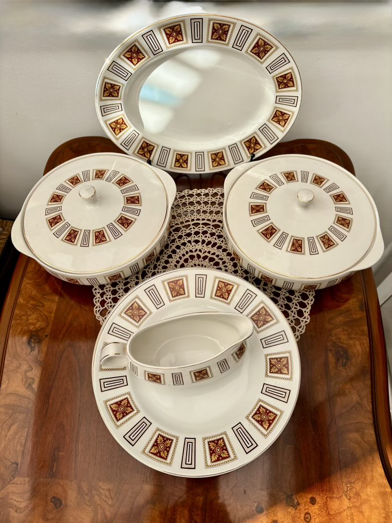 Vintage bone china dinner service set, top view, including two lidded serving tureens, an oval serving platter, a gravy boat, and a dinner plate, all decorated with a geometric border of dark red and gold diamond motifs with black rectangular frames, displayed on a wooden sideboard with a lace doily