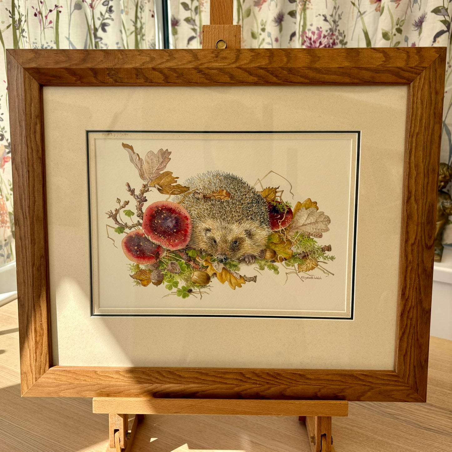 Framed artwork of a hedgehog among flowers and leaves on a wooden easel.