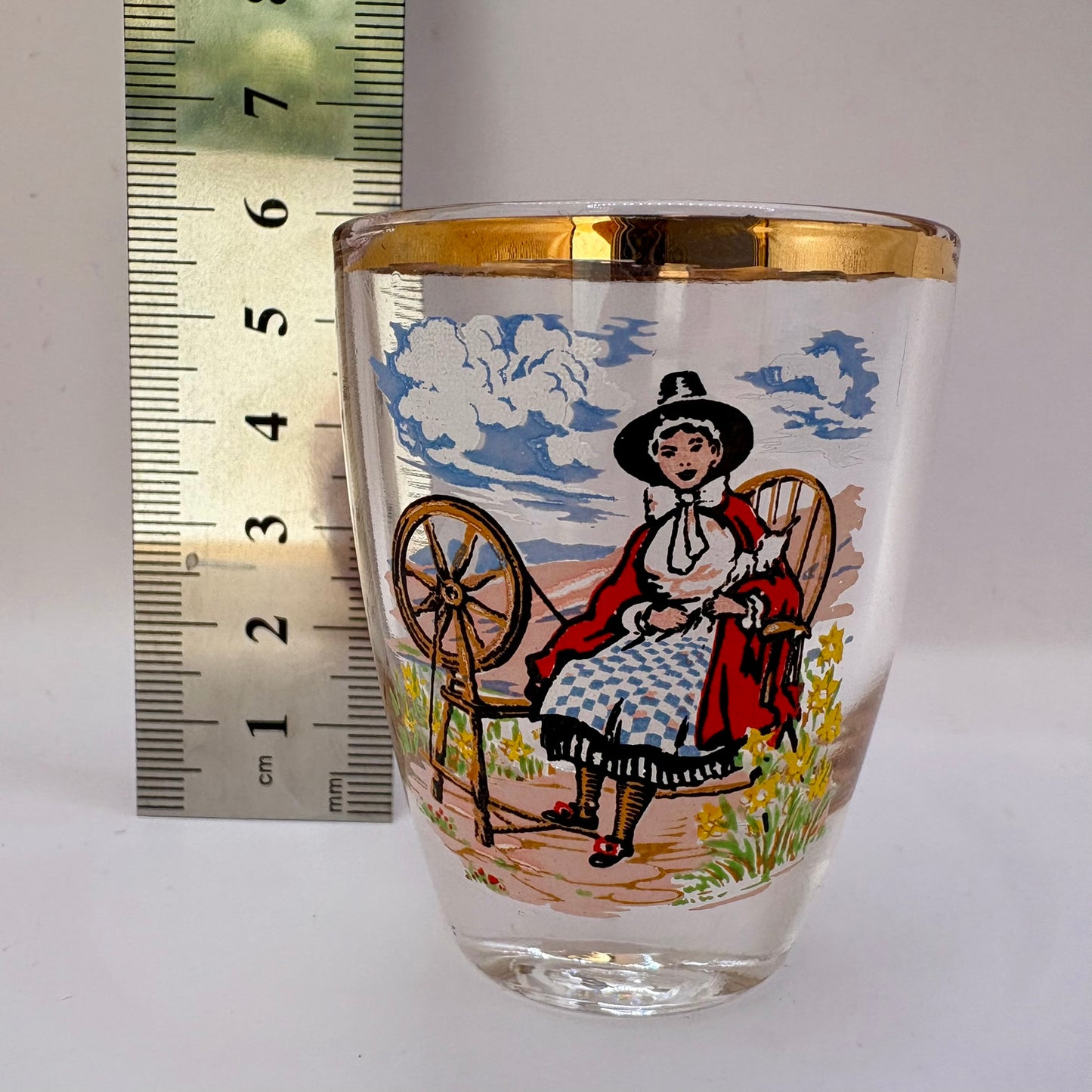 Decorative glass with a printed design of a woman spinning, next to a ruler for scale.