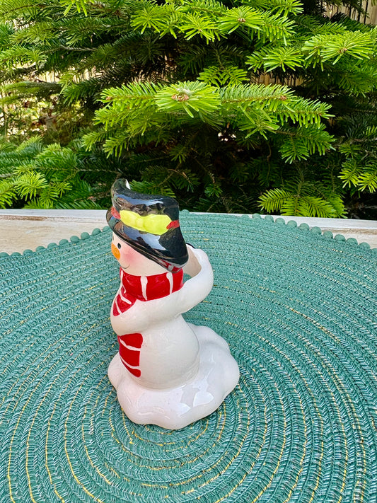 Decorative snowman figurine with a red scarf on a green textured surface with a Christmas tree in the background.