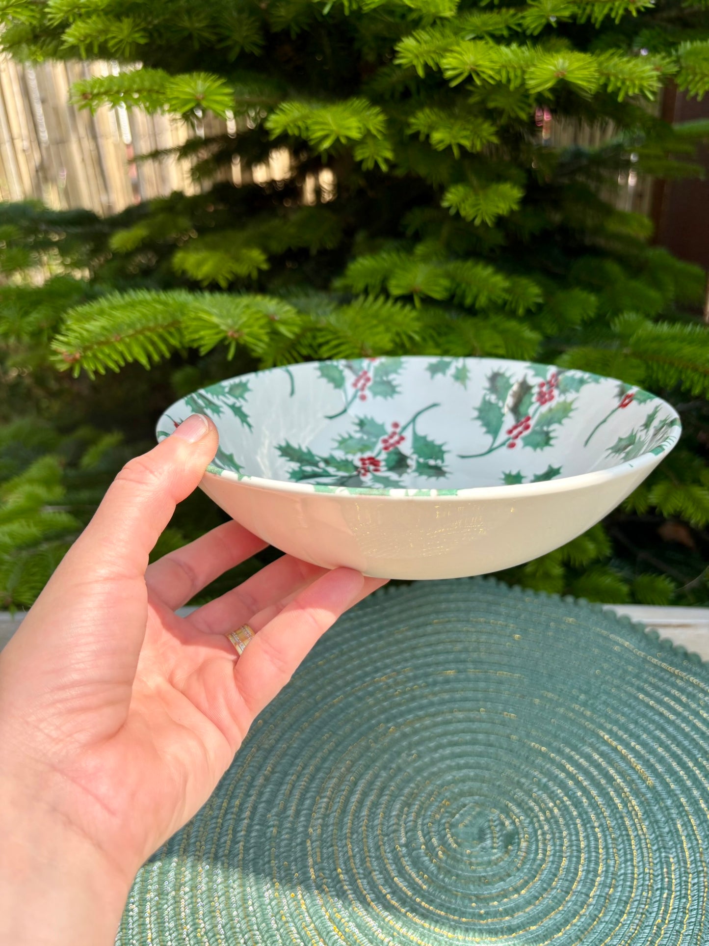 Hand holding a small decorative bowl with holly design in front of a Christmas tree.