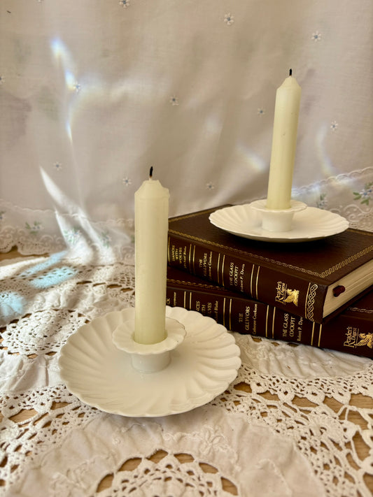 Two white candles in decorative holders on a lace tablecloth with books in the background.