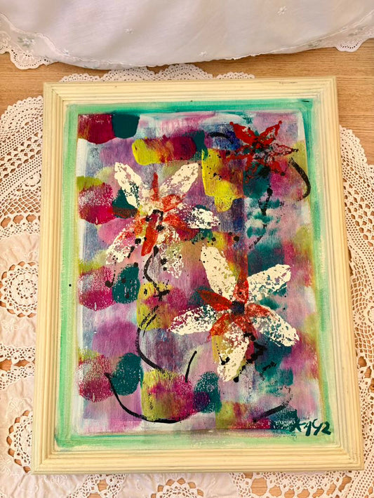 Colorful abstract painting of flowers in a light wooden frame on a lace tablecloth.