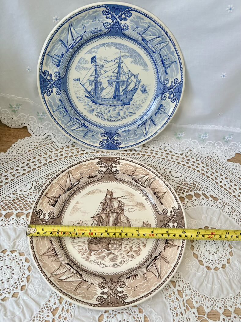 Two decorative plates with ship designs on a lace tablecloth, measured with a tape measure.