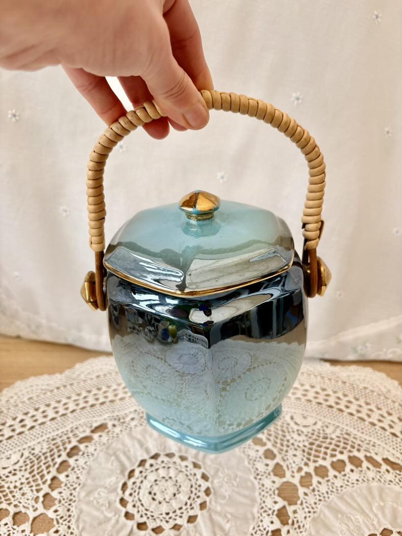 Blue ceramic jar with a lid and woven handle held by a hand on a lace tablecloth.