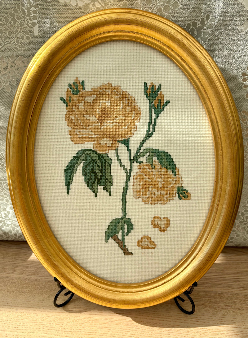 A vintage oval framed needlepoint picture featuring a yellow rose with green leaves on a neutral background, set in a gold-colored frame.