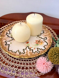 Two white candles on a decorative plate with a doily underneath, surrounded by flowers.
