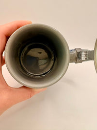Person holding a ceramic mug with a metal handle against a plain background
