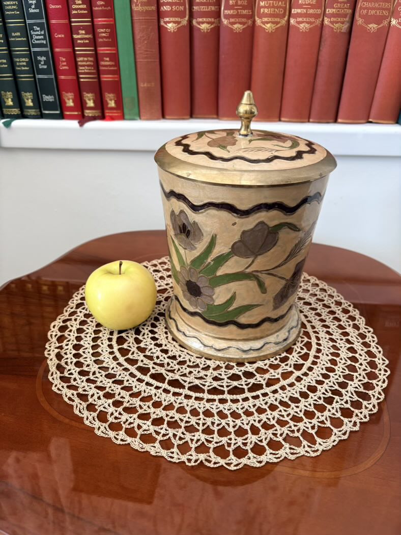 Decorative ceramic jar with floral design on a doily, apple, and books in the background