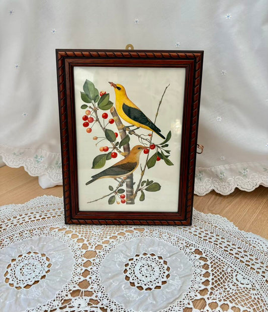 Framed artwork of birds on a branch with berries on a lace tablecloth.