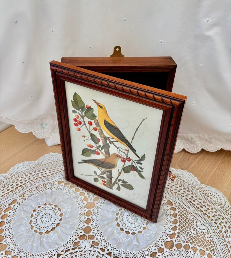 Framed artwork of birds on a branch with berries on a lace tablecloth.