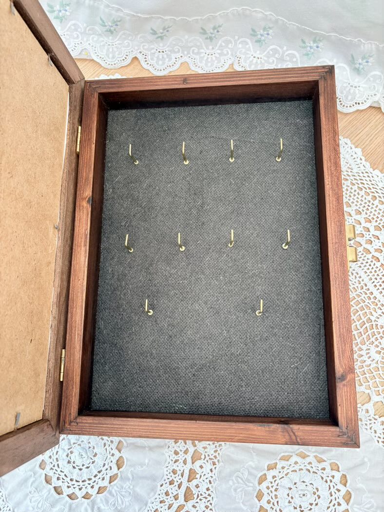 Wooden keys box with gray interior displaying gold hooks on a lace tablecloth.
