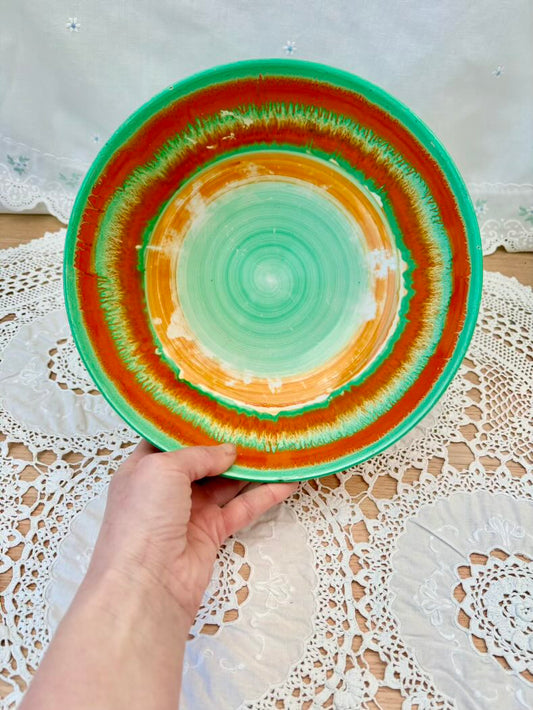 Hand holding a ceramic plate with green and orange concentric circles on a lace tablecloth.