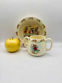 Children's ceramic cup and plate set with cartoon characters, accompanied by a yellow apple on a white background.