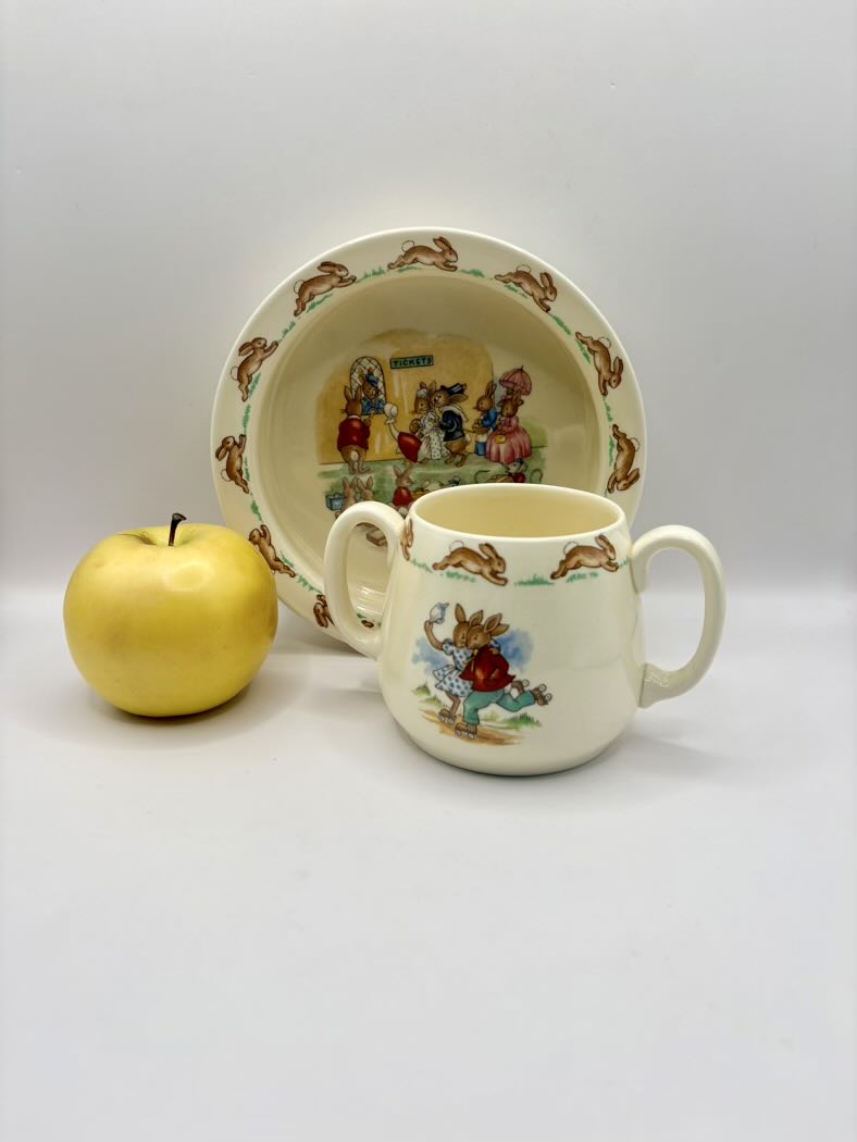 Children's ceramic cup and plate set with cartoon characters, accompanied by a yellow apple on a white background.