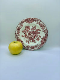 Decorative plate with intricate red patterns next to a yellow apple on a light gray background
