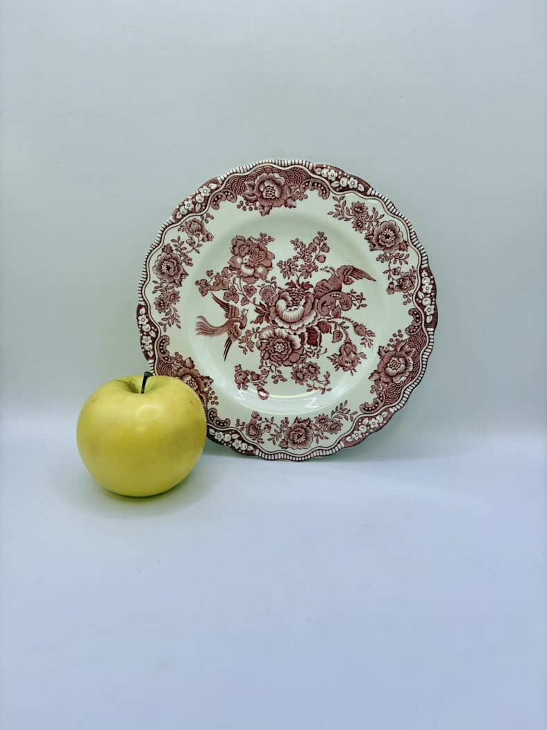 Decorative plate with intricate red patterns next to a yellow apple on a light gray background