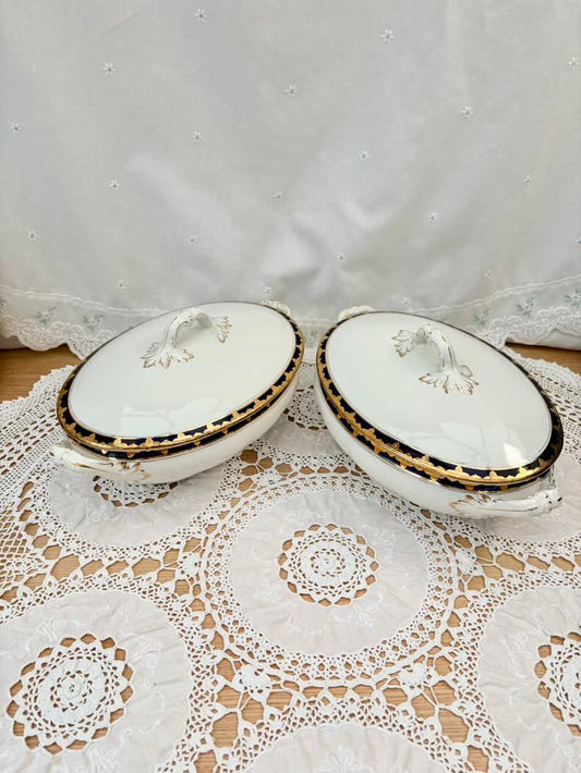 Two white ceramic covered dishes with gold accents on a lace tablecloth.