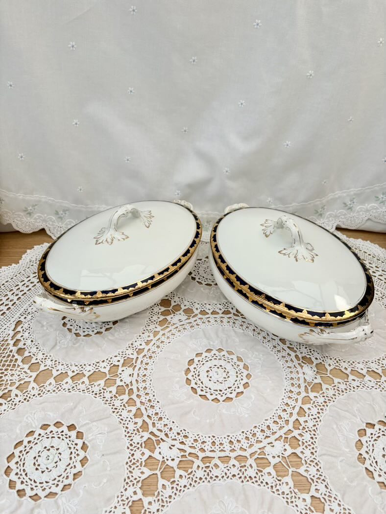 Two white ceramic covered dishes with gold accents on a lace tablecloth.