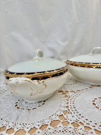 Two ceramic covered dishes with gold accents on a lace tablecloth.