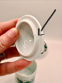 Hand holding a white ceramic container with a lid, showing a small amount of liquid inside.