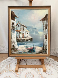 Framed painting of a coastal scene with houses and a boat on an easel.