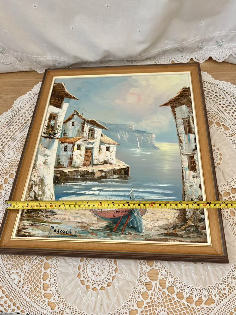 Framed painting of a coastal scene with houses and a boat, placed on a lace tablecloth.