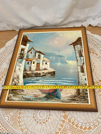 Framed painting of a coastal scene with houses and a boat, placed on a lace tablecloth.