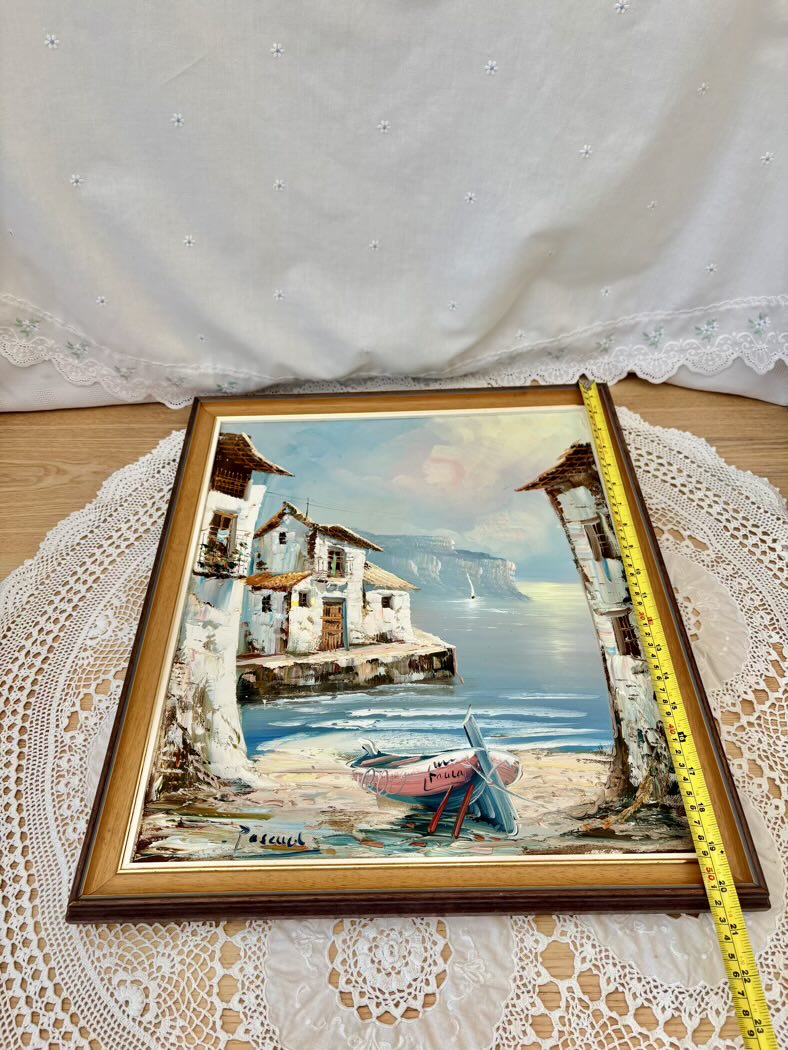 Framed painting of a coastal scene with houses and a boat on a lace doily.