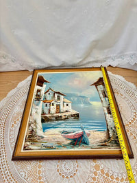 Framed painting of a coastal scene with houses and a boat on a lace doily.
