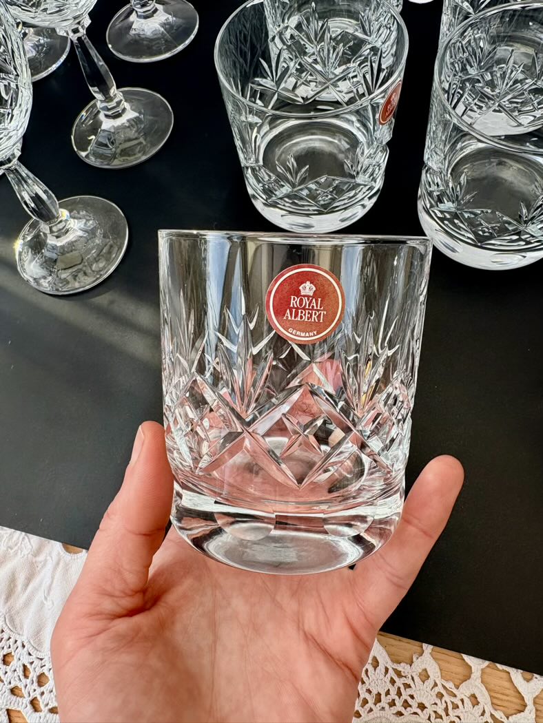 Hand holding a crystal glass with 'The Royal Albert' logo on a dark surface with other glasses.