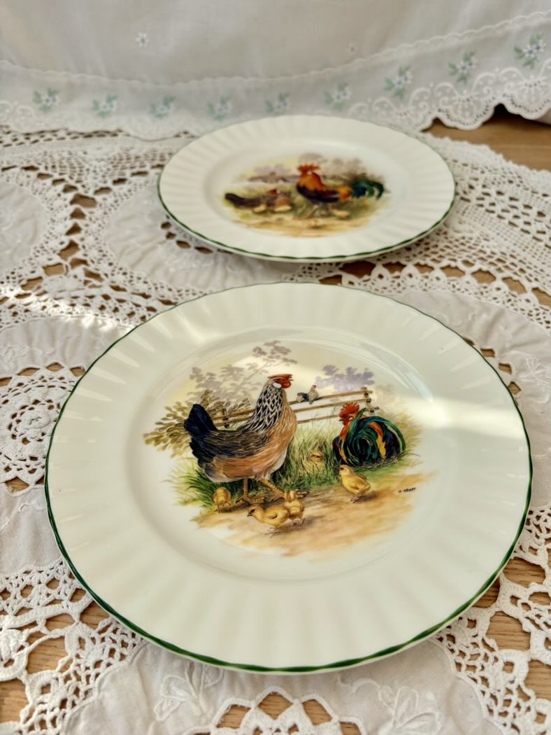 Two decorative plates with rooster designs on a lace tablecloth.
