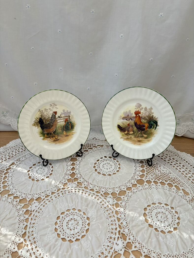 Two decorative plates with rooster designs on a lace tablecloth.