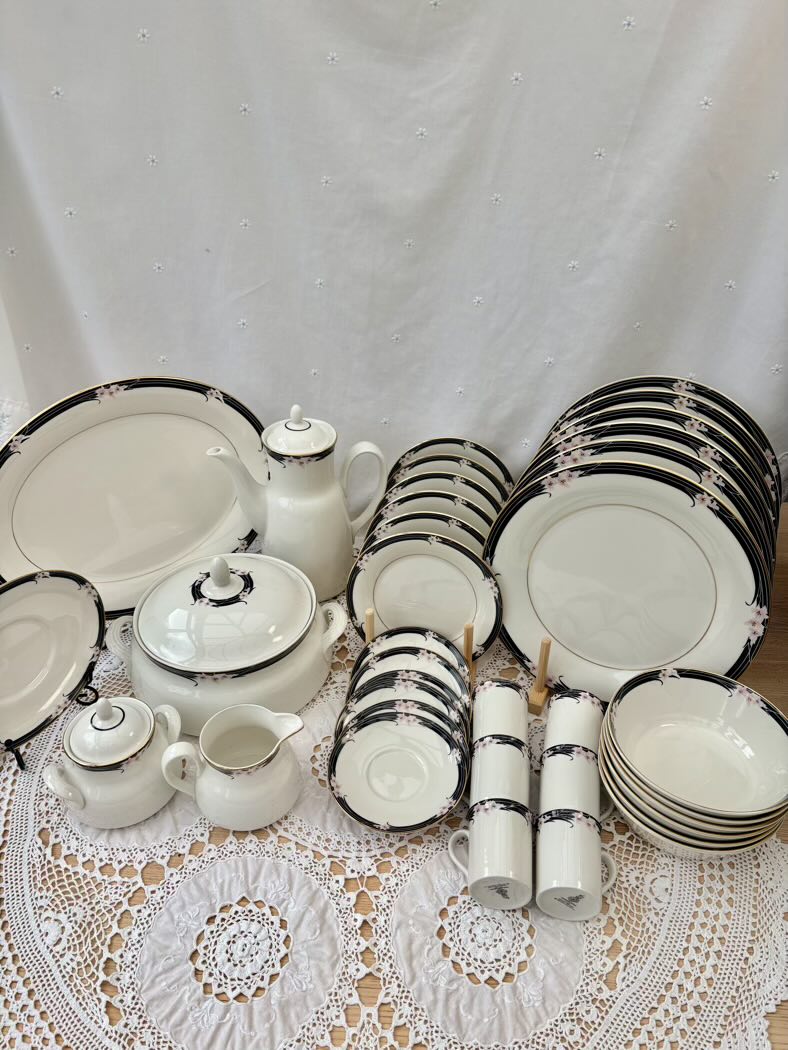 Set of white ceramic dinnerware with black trim on a lace tablecloth.
