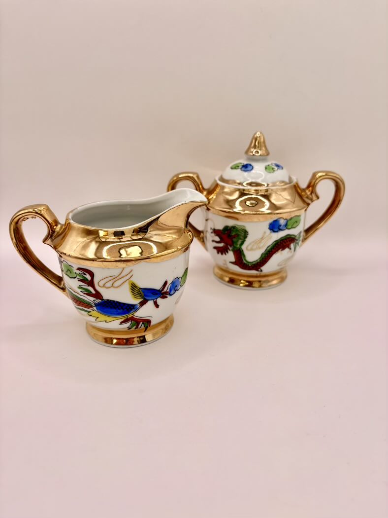 Decorative ceramic creamer and sugar bowl with gold accents and colorful designs on a light pink background.