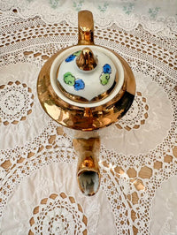 Decorative teapot with a white shade featuring colorful floral patterns on a lace tablecloth.