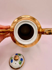 Gold and white ceramic teapot with a hand holding it on a light pink background