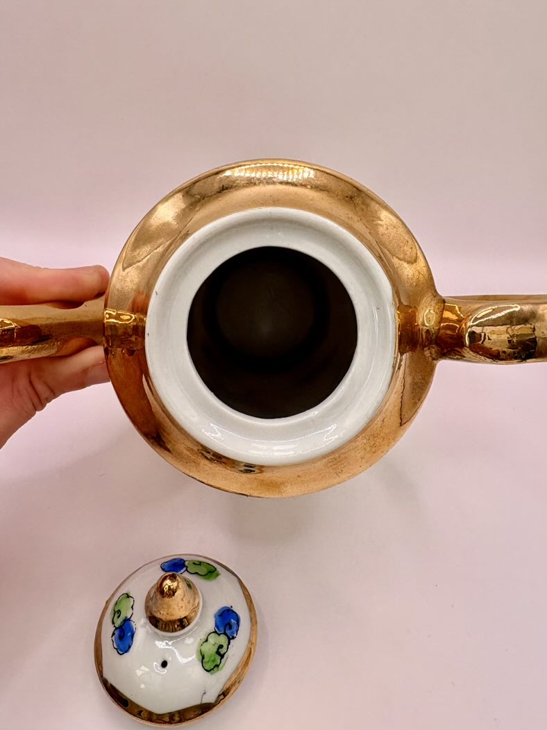 Gold and white ceramic teapot with a hand holding it on a light pink background