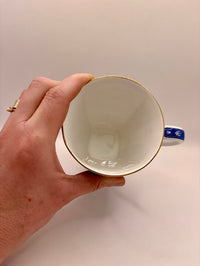 Hand holding a ceramic cup with gold rim against a plain background