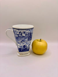Blue and white mug with architectural design next to a yellow apple on a light gray background