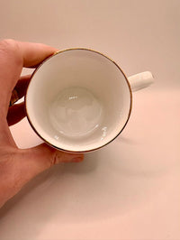 White mug with gold rim held by a hand on a beige background