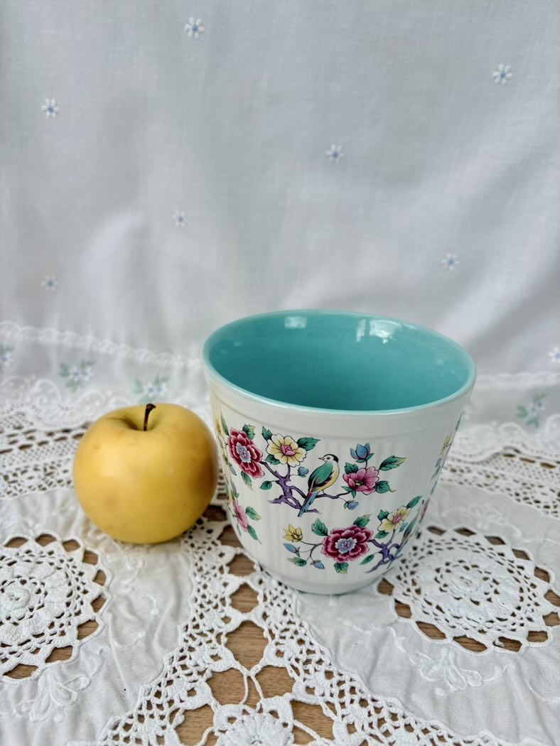 Decorative ceramic cup with floral patterns next to an apple on a lace tablecloth.