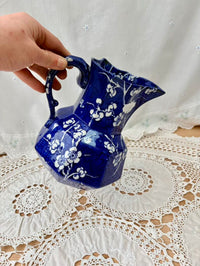 Blue ceramic pitcher with white floral patterns held by a hand on a lace tablecloth.