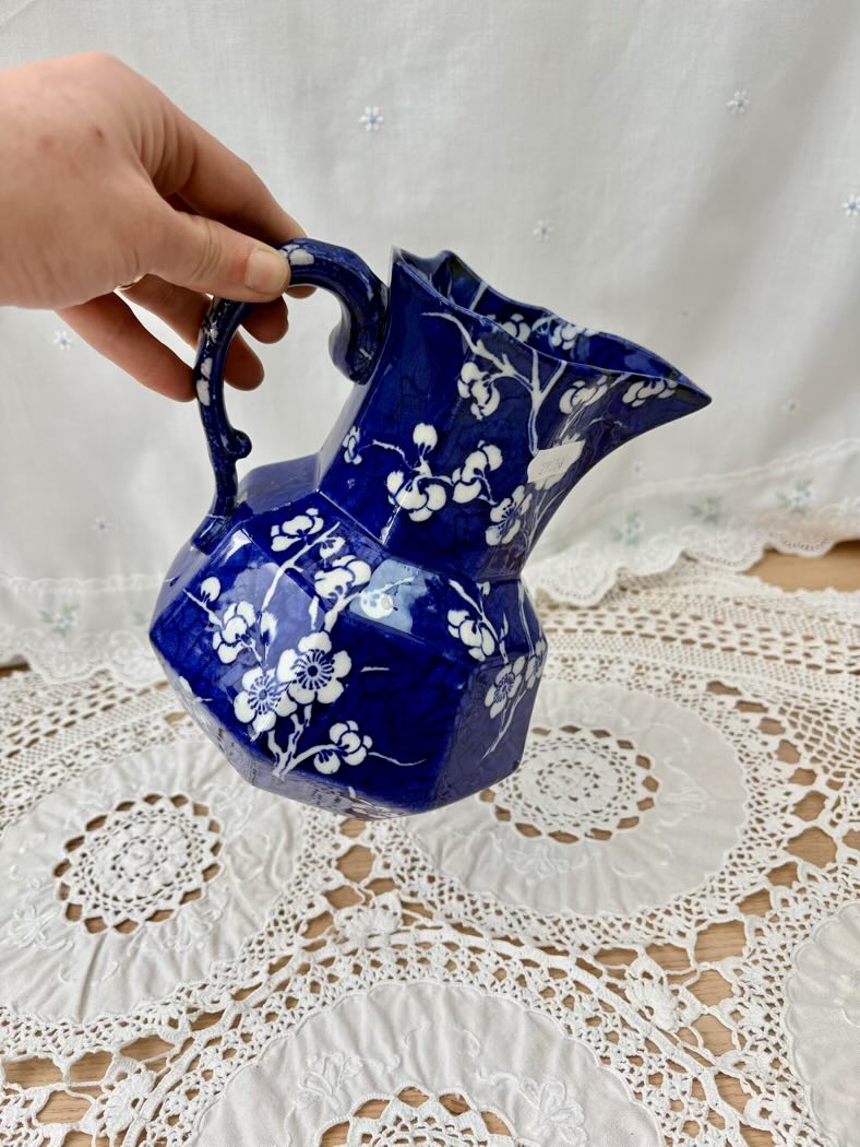 Blue ceramic pitcher with white floral patterns held by a hand on a lace tablecloth.