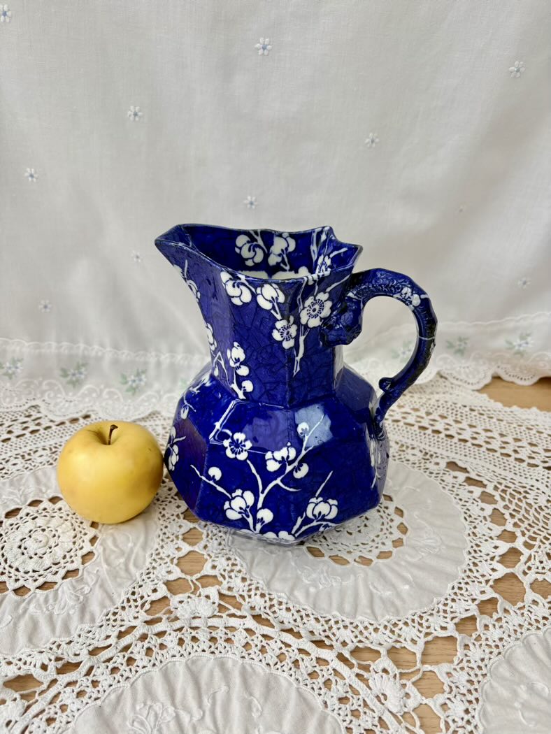 Blue ceramic pitcher with white floral patterns on a lace tablecloth with an apple beside it.