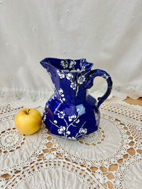 Blue ceramic pitcher with white floral patterns on a lace tablecloth with an apple beside it.