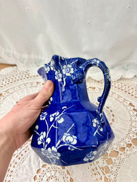 Blue glass pitcher with white floral patterns held by a hand on a lace tablecloth.